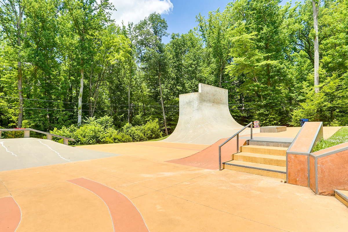 A sunny skate park with multiple concrete ramps, metal railings, and a staircase. Beyond the park is a forested area.