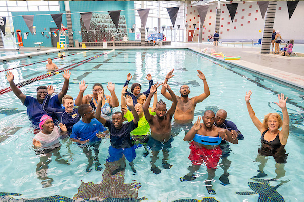 A group of disabled young adults poses together in an indoor pool, all with their arms raised.
