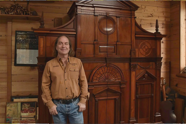 A man posing next to a large and ornately carved wooden cabinet or armoire.