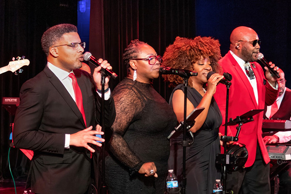 Four adults in formalwear sing into microphones on a stage, with a backing band behind them.
