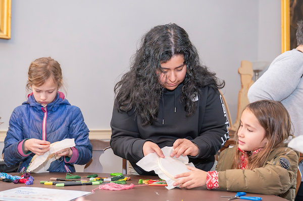 At a table, two kids and an adult work on sewing projects using cloth, embroidery hoops, and colorful embroidery floss.