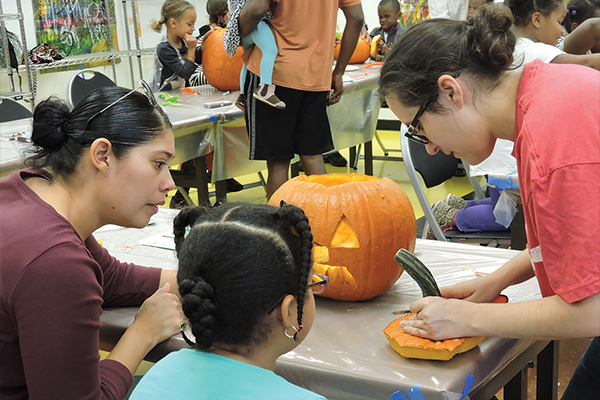 People sit at plastic-covered tables carving and decorating pumpkins.