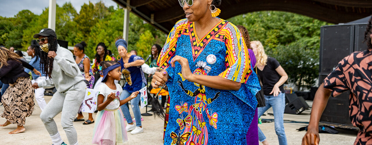 Festival attendees dance together in front of an outdoor stage, including a person in a bright blue patterned garment and a child in a pastel dress.