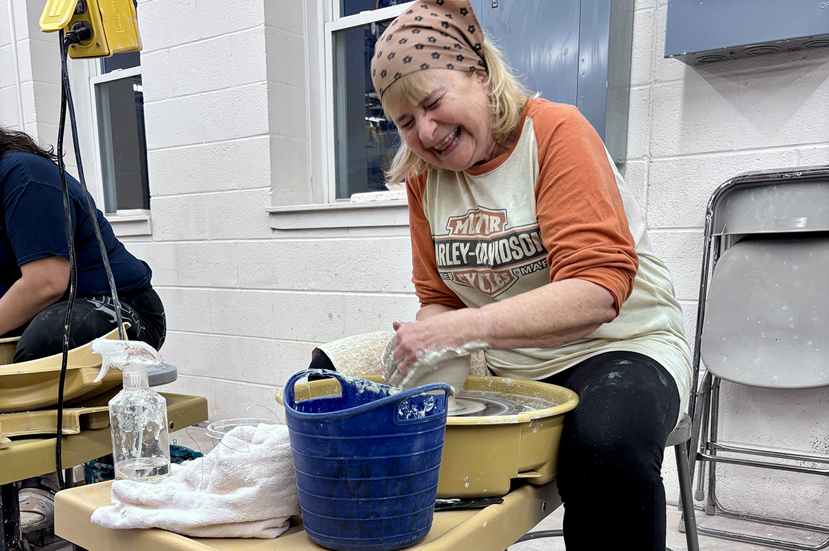 A laughing woman with a scarf on her head sits at a pottery wheel, shaping clay into a bowl.