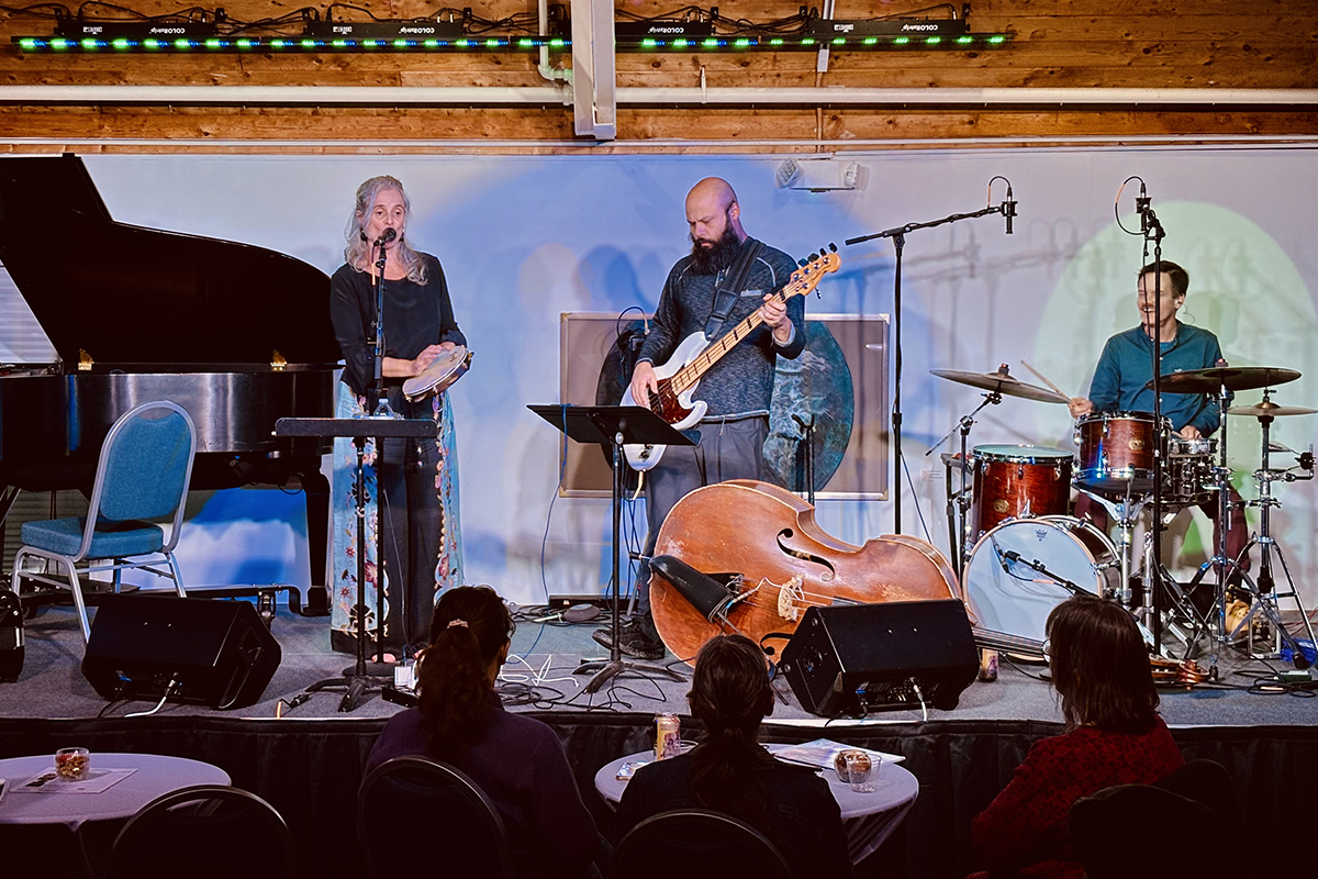 Three musicians perform on a stage in front of a seated audience. They include a singer/tambourine player, a drummer, and a bass guitarist.
