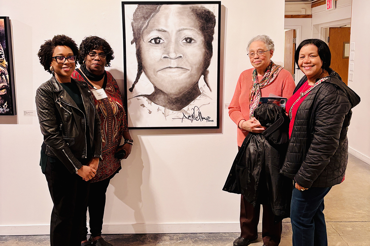 Four smiling people pose in an art gallery next to a large, framed, black-and-white portrait of a young child.