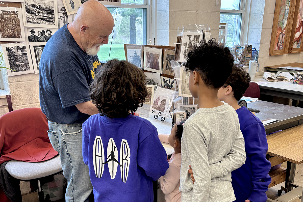 A resident artist talks to four young visitors in his studio, showing them small prints and etchings.