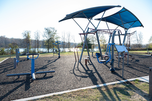 Wide view of an outdoor fitness area featuring multiple exercise stations, shade canopies, and a lake in the background.