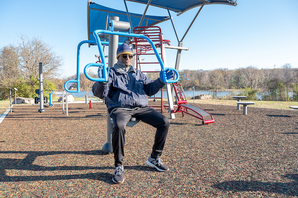 Person seated on an outdoor chest press machine beneath a shade canopy at a lakeside fitness park.