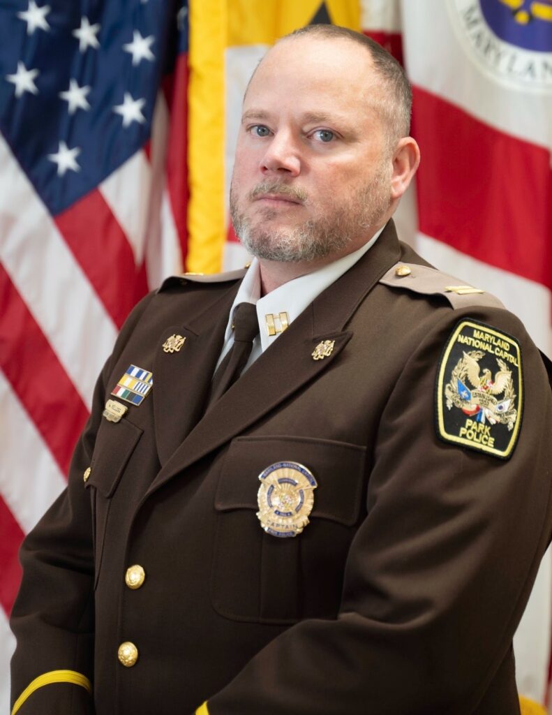Uniformed park police official in dress uniform, photographed before American, Maryland, and Prince George's County flags.