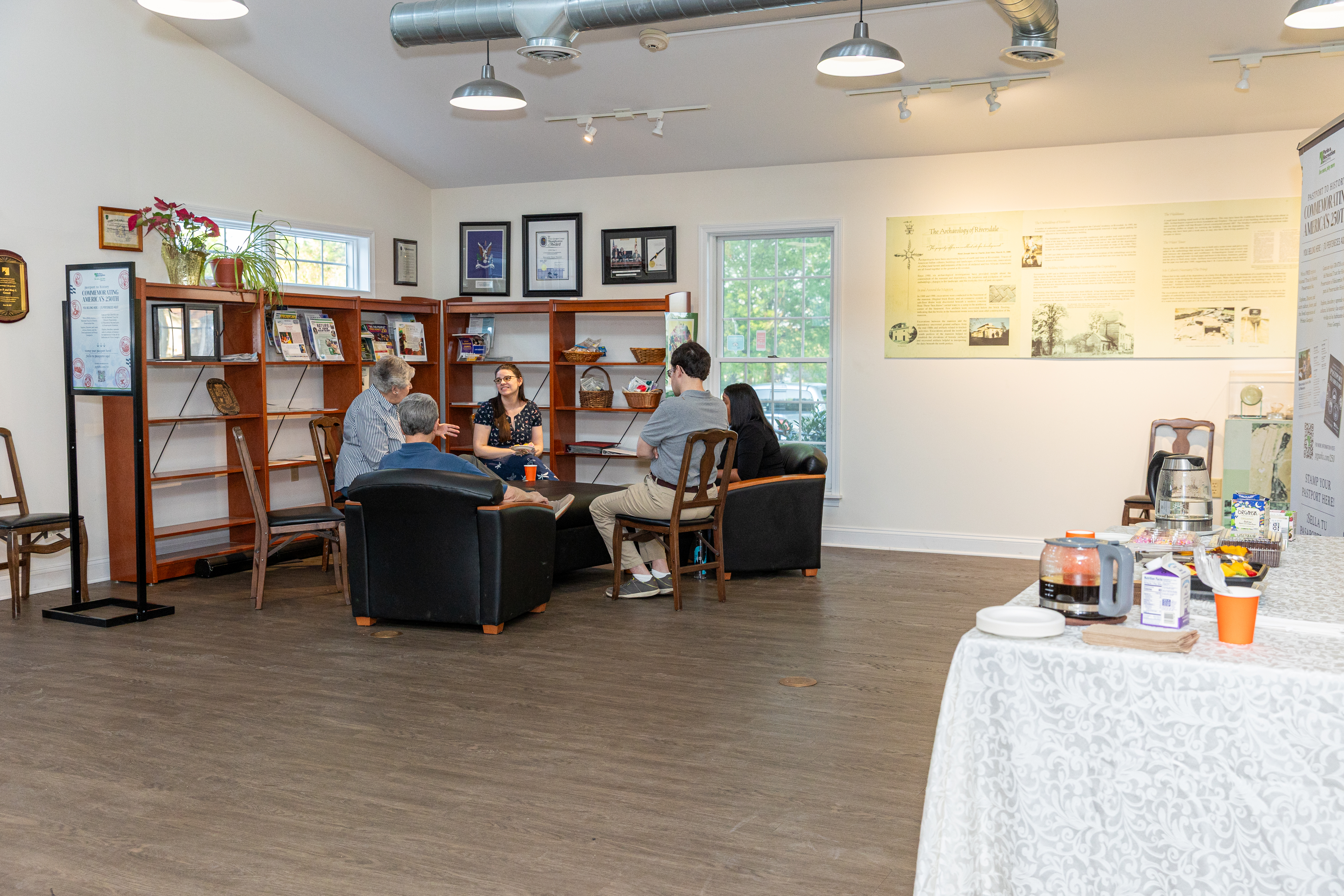 Community members seated in a museum gallery for a small group discussion, with bookshelves, exhibits, and a refreshment table nearby.