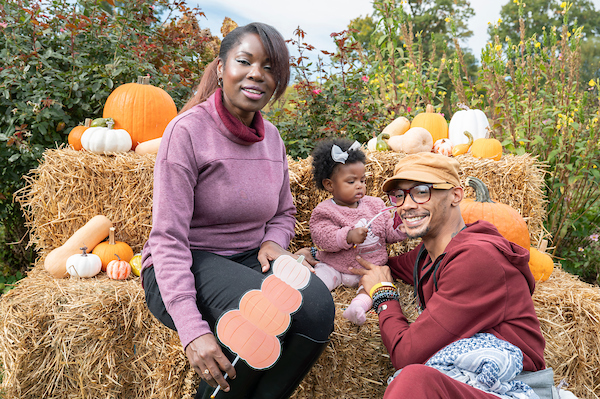 Family with a young child sitting on hay bales among pumpkins and gourds at an outdoor harvest festival.