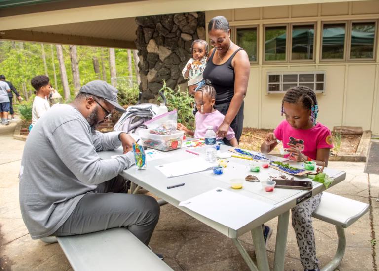 Families and children gather under a canopy tent, creating arts and crafts together during an outdoor community event at a nature center.