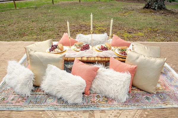 A picnic assortment of pink, gold and white pillows around a table with food, candles, and decorations all on a carpet in the grass.